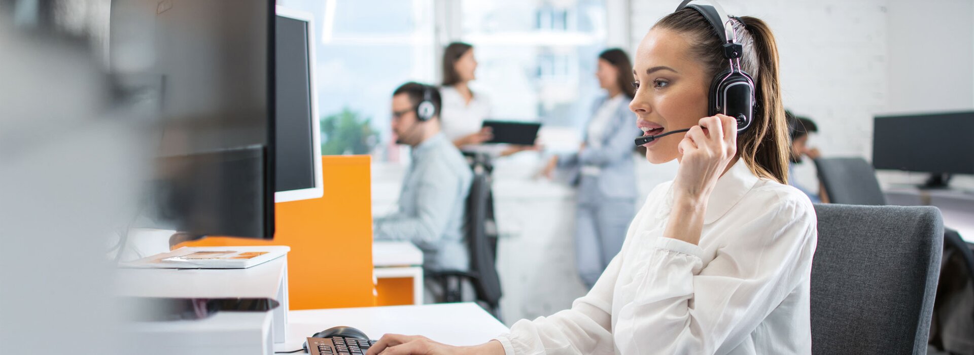 Kundendienstmitarbeiterin mit Headset im Büro Frau mit Headset arbeitet am Computer im modernen Büro mit Kollegen im Hintergrund